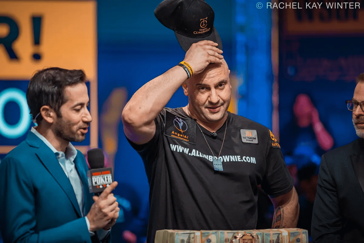 Michael Mizrachi takes off his cap after winning the 2025 WSOP Main Event, with the championship bracelet and prize money in front of him, joined by a reporter and WSOP official.